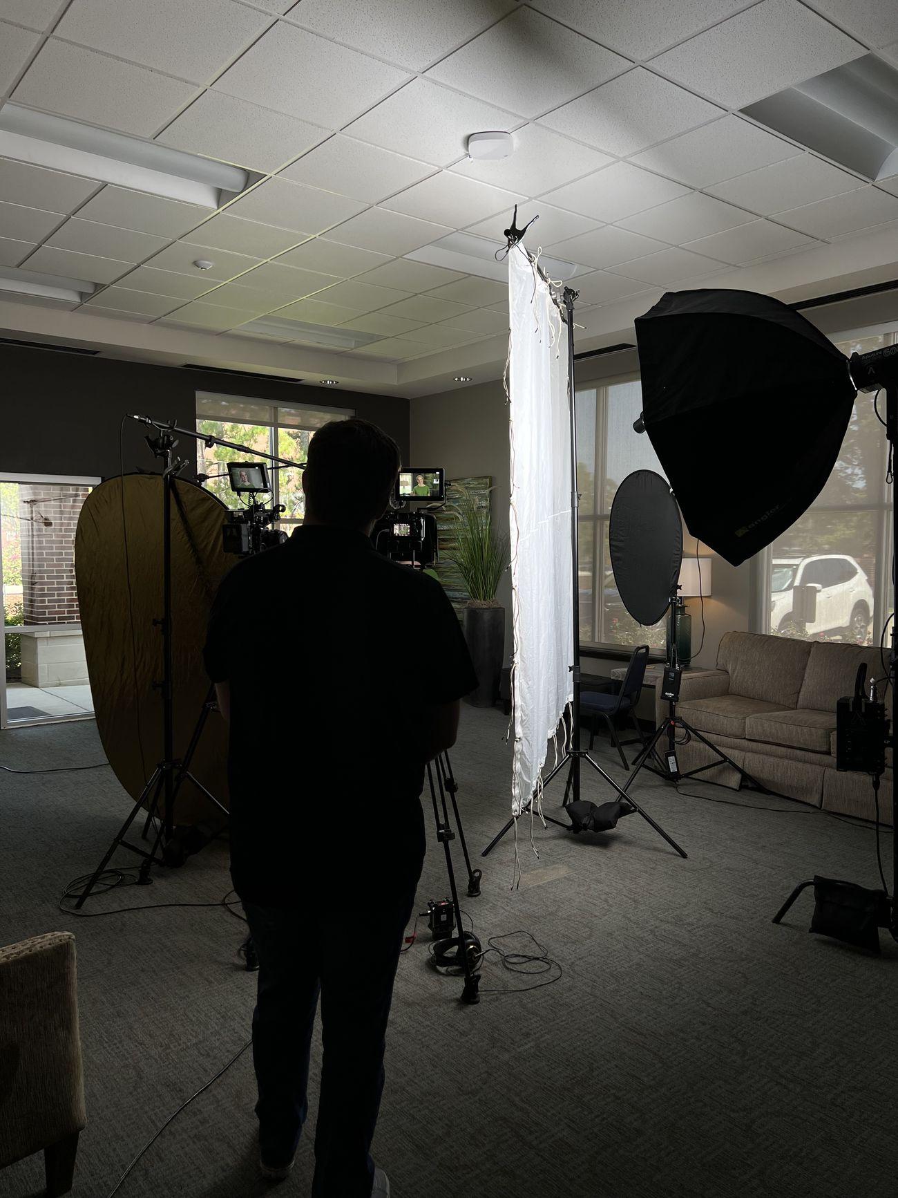 A video production lighting and camera setup in an office, with the operator silhouetted against softboxes and diffusion